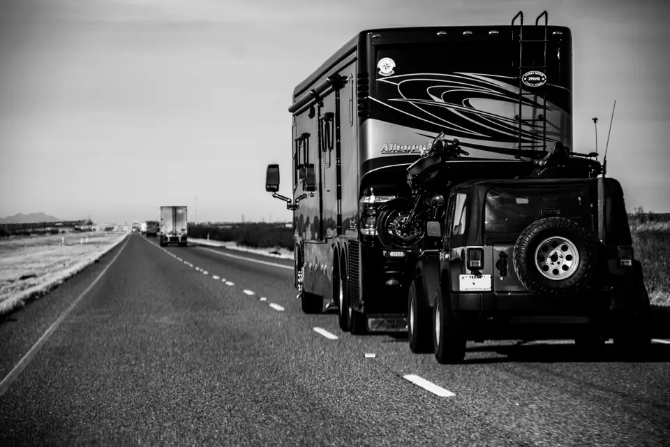 RV drives down the road pulling a jeep behind it. Their FRVA membership plaque can be seen hanging from their RV ladder. Ahead of them on the highway, semi-trucks drive in the same direction on this two-lane road.