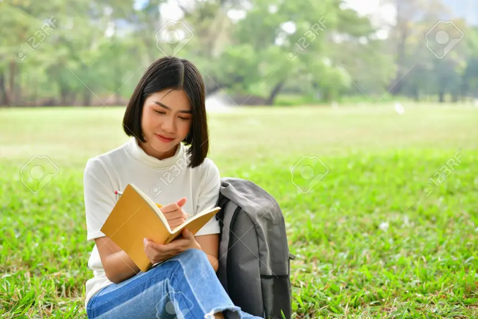 young asian woman sitting outside on the grass reading with a gray backpack beside her