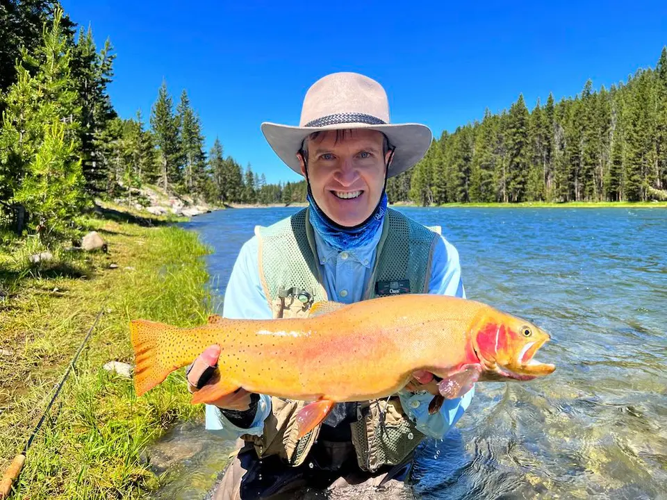A photo of John Healey holding a large cutthroat trout under a bright blue sky next to a river on the About page of The Concise Verse.